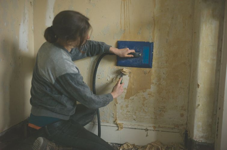 Young woman removing wallpaper with scraper and steamer while crouching